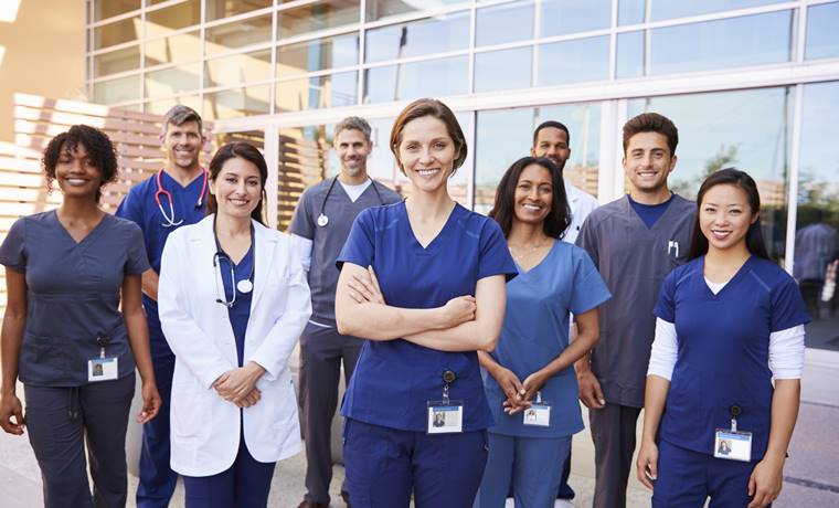 A group of health care professionals, doctors and nurses, standing outside together looking at the camera and smiling.