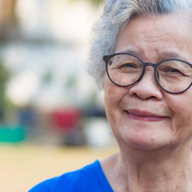 A portrait of an elderly woman with short white hair wearing glasses smiling and looking at the camera while standing in a garden of a senior care facility.