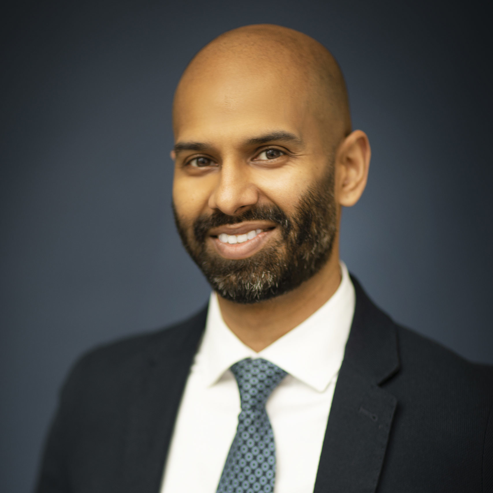 Photo of Shaun Dias wearing a suit and tie in front of a black background
